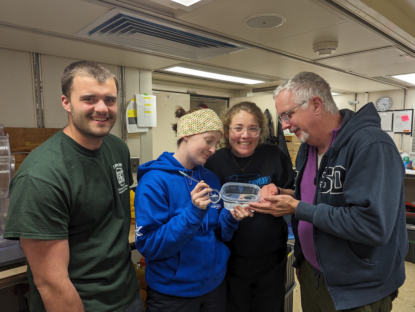 four people standing in a lab and adoringly looking at a tupperware container with a small baby octopus in it