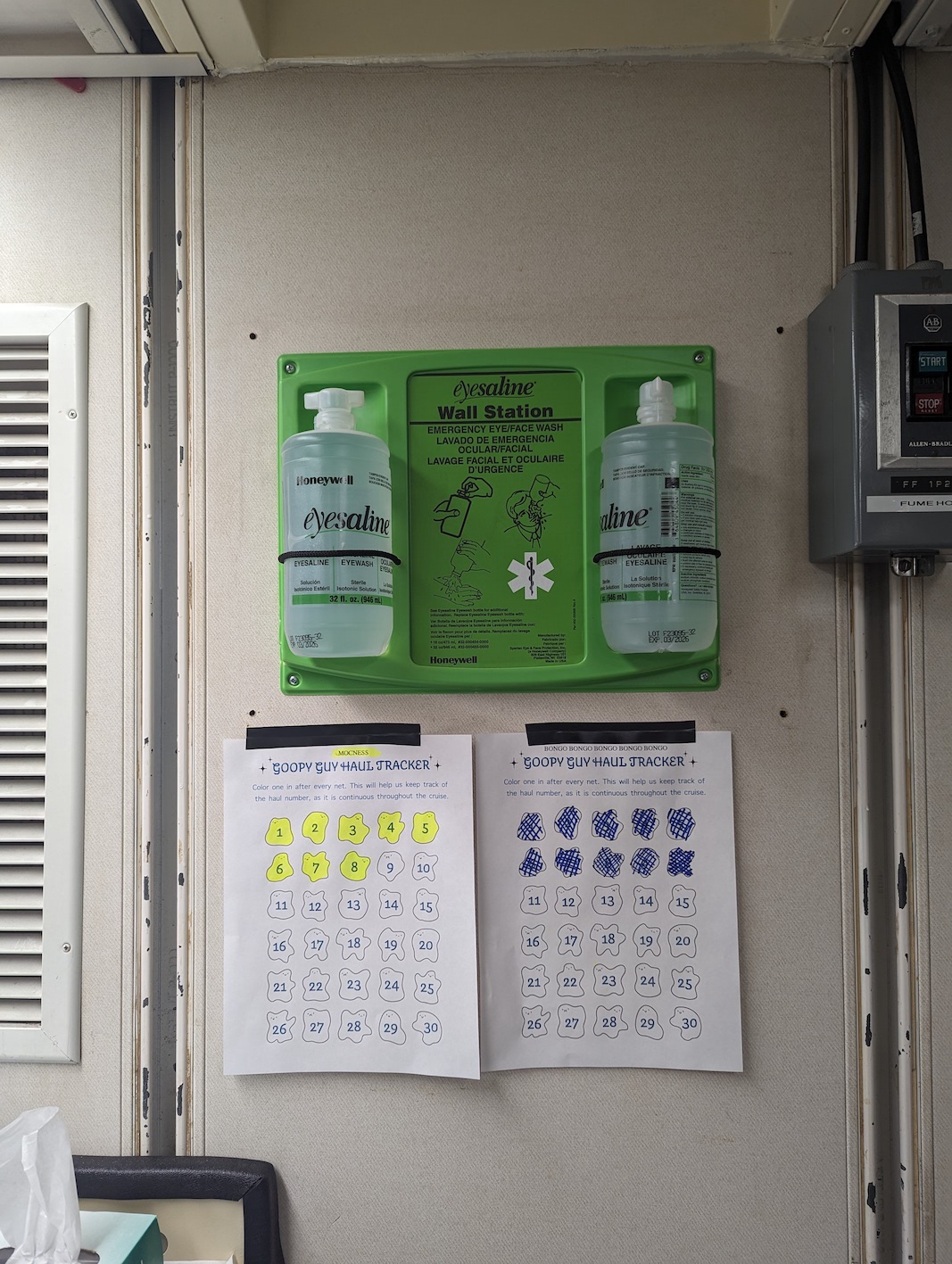 image of wall in lab, with eye wash station and two coloring sheets titled 'gooey guys' for tracking the net hauls